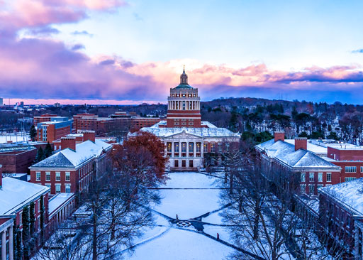 Snow-covered University of Rochester campus with the Rush Rhees Library as the central focal point, framed by academic buildings and winter trees.