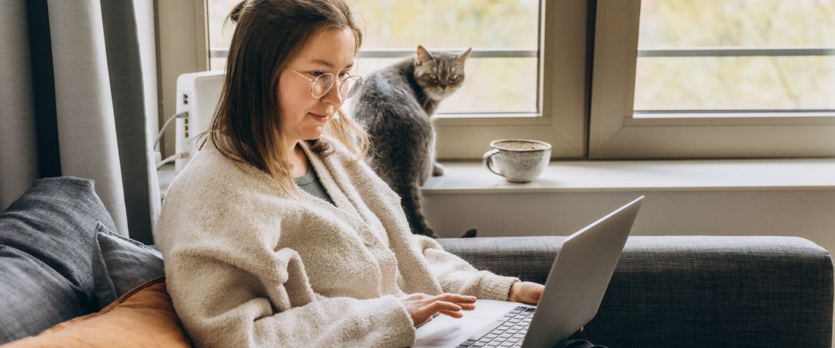 A woman sits on a couch with a laptop, while a cat gazes out the window, creating a cozy indoor scene.