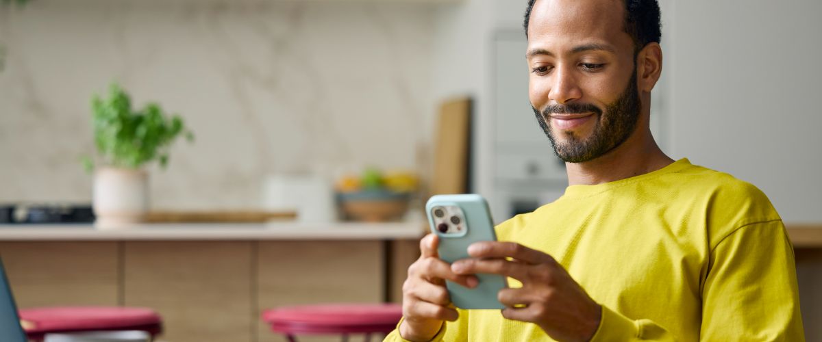 A man wearing a yellow shirt is engaged with his smartphone, focused on the screen in a casual setting.