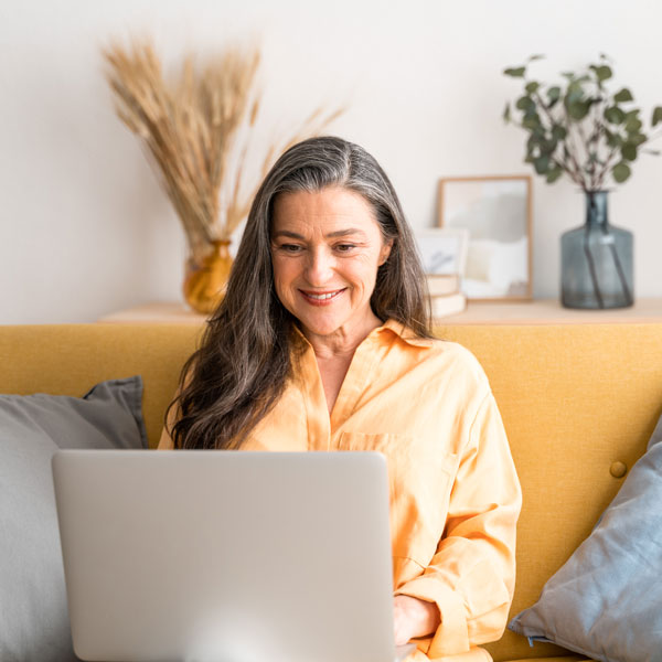 A woman sitting on a couch, focused on her laptop, with a cozy living room setting in the background.