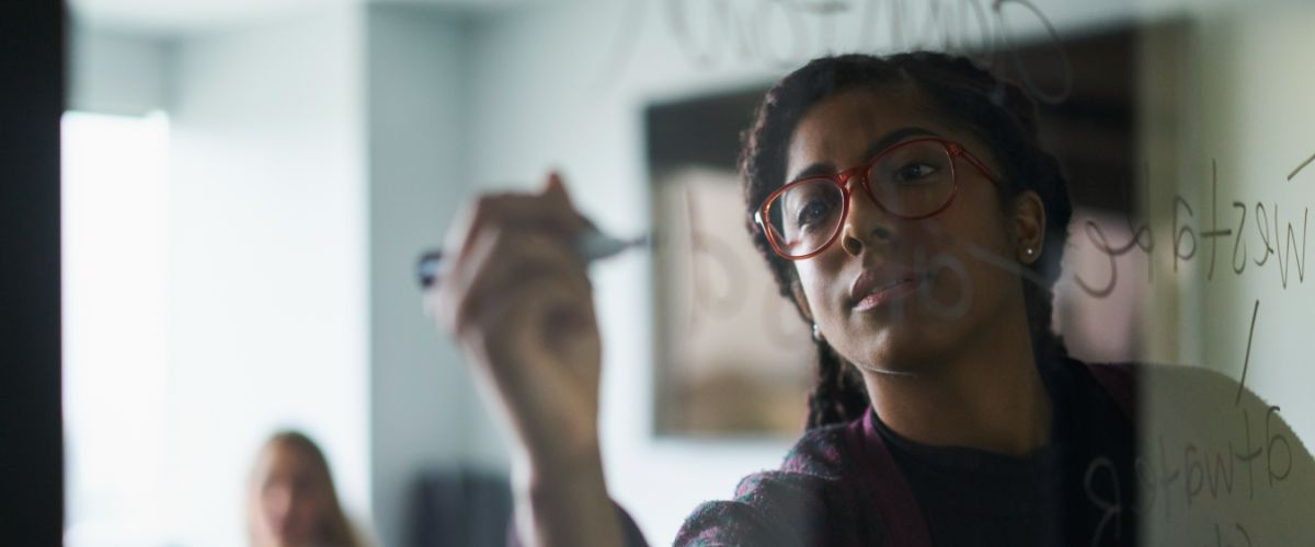 A person with red glasses writing on a clear glass board.