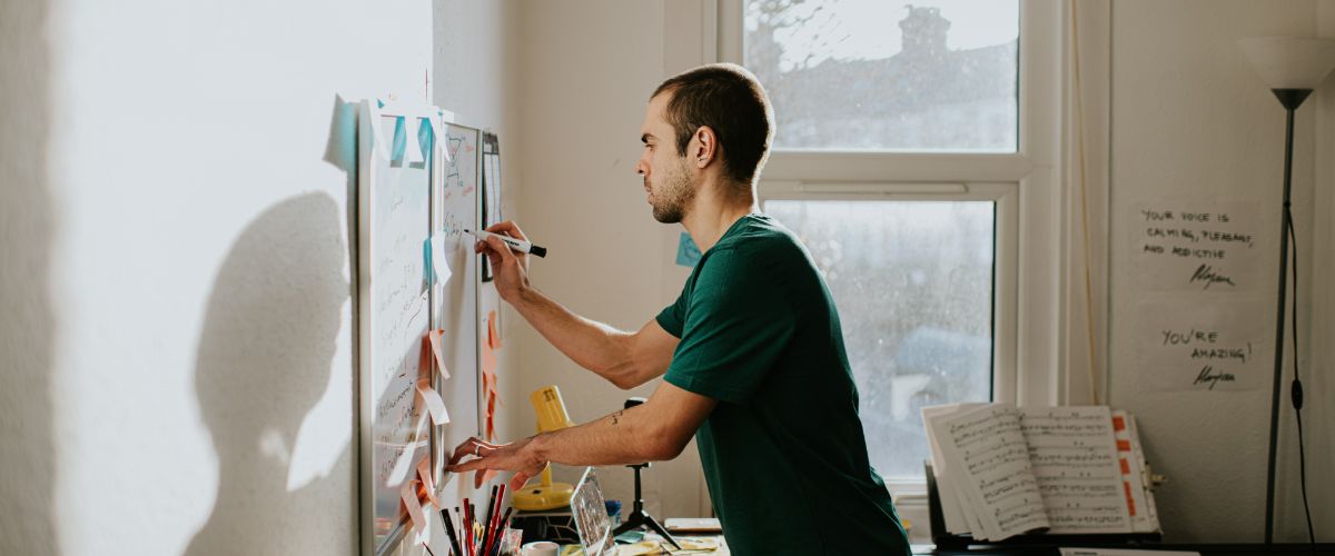 Man writing on a whiteboard covered with sticky notes in a sunlit room.